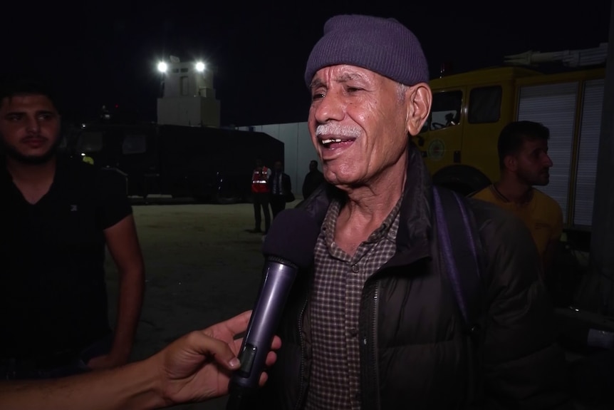 An older man with white moustache and a purple beanie speaks at night time, while someone holds a microphone to his face