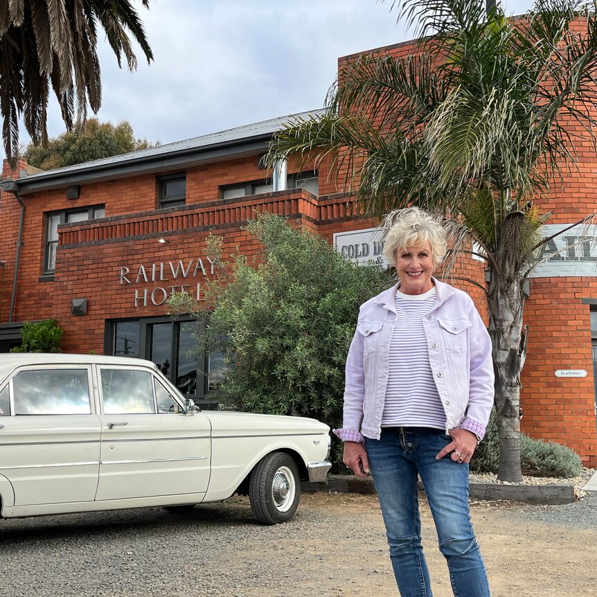 Woman standing and smiling outside a red brick pub named "Railway Hotel" with vintage car in background. 