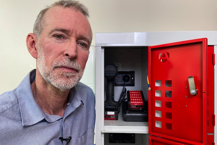 a man poses next to a metal locker with a red door, inside it is a powerpoint with a battery charger