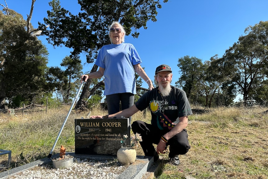 A woman in a blue tshirt holds a walking stick with her hand on the shoulder of a man kneeling by a headstone. 