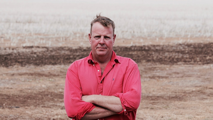 Grain grower Adrian Lyons stands in front of a barren-looking field with his arms crossed.