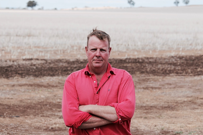 Grain grower Adrian Lyons stands in front of a barren-looking field with his arms crossed.