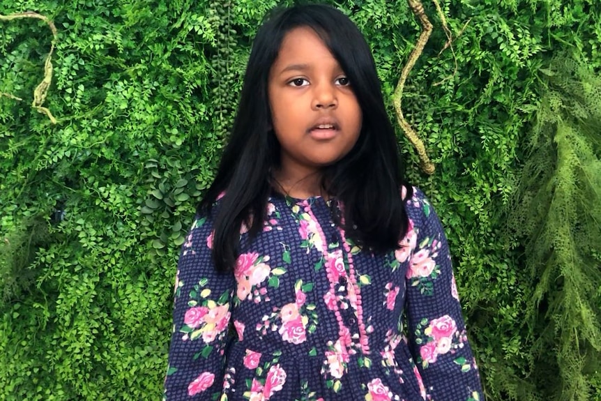 A child with dark hair stands in front of a hedge.