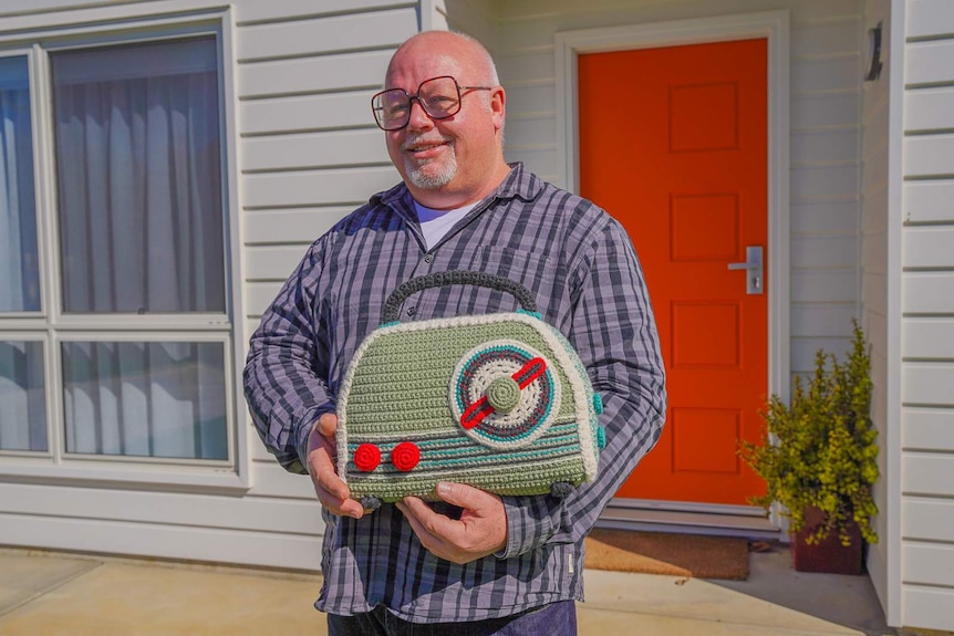 A man in large glasses smiles outside a white wood panelled home holding a life-sized crocheted radio.