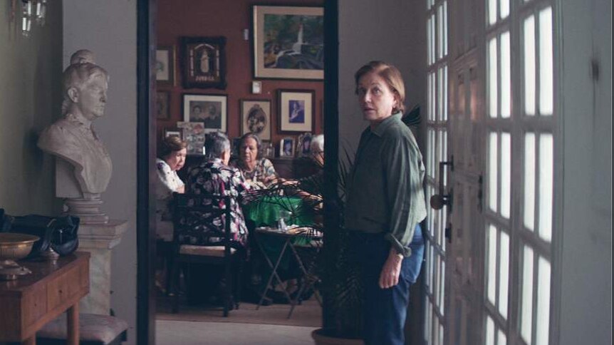 60ish woman in slacks and shirt stands in hallway of upper class apartment with marble busts and paintings on walls.