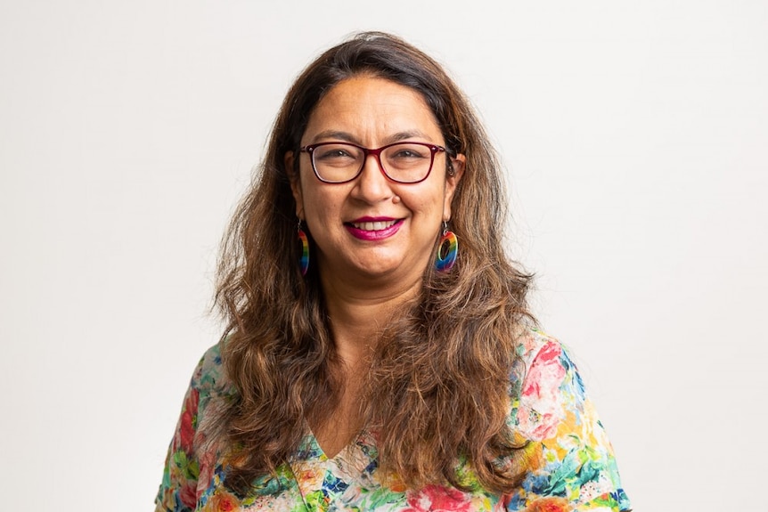 A woman with glasses, long brown hair and a multi-coloured shirt smiles at the camera
