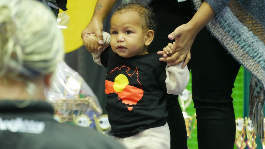 An Aboriginal baby stands with the help of his mum wearing a black T-shirt with the Aboriginal flag on it.