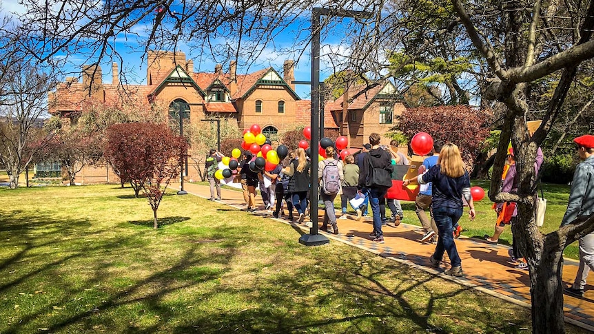Students carrying red, yellow and black balloons walk in a line towards UNE's Boolomimbah building