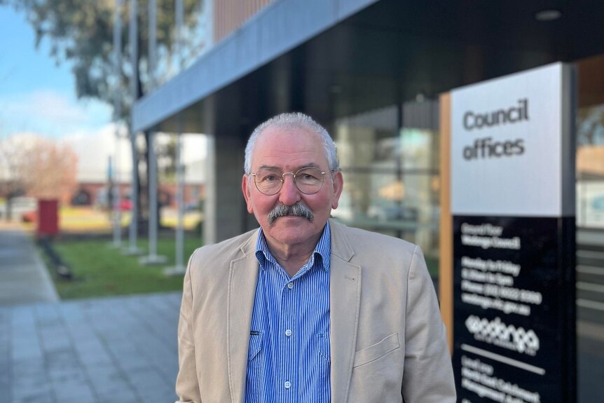 older man with glasses and moustache looks directly at the camera, council offices signage can be seen in the background.