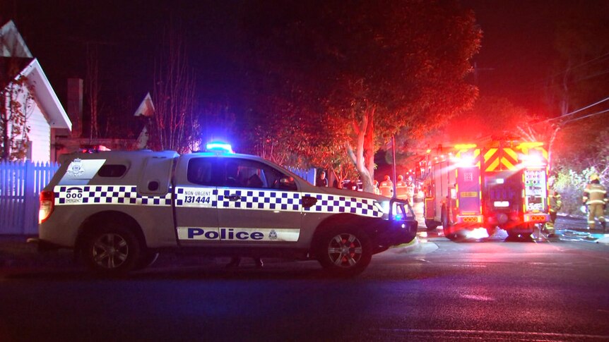 A police car and fire engine with emergency lights activated are parked in a dark street outside a white brick home