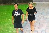 A man in sportswear and a woman in shorts and a blouse walk walk down a path near a car park.