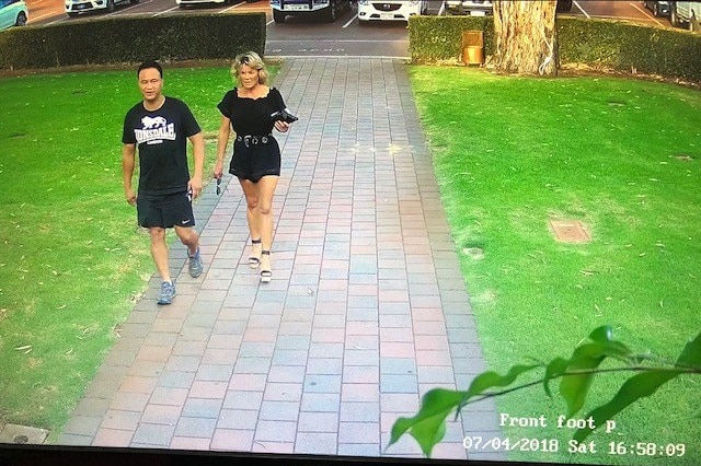 A man in sportswear and a woman in shorts and a blouse walk walk down a path near a car park.