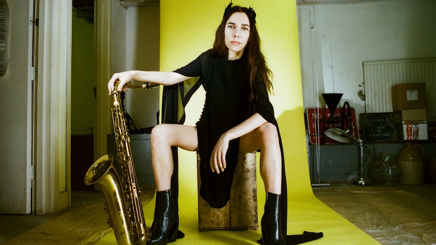 PJ Harvey poses with a saxophone in front of a yellow backdrop.