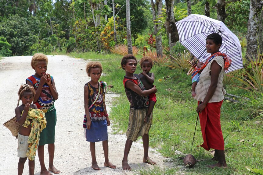 A woman with an umbrella and several young children stand by a dirt road. The woman and one child are holding babies.