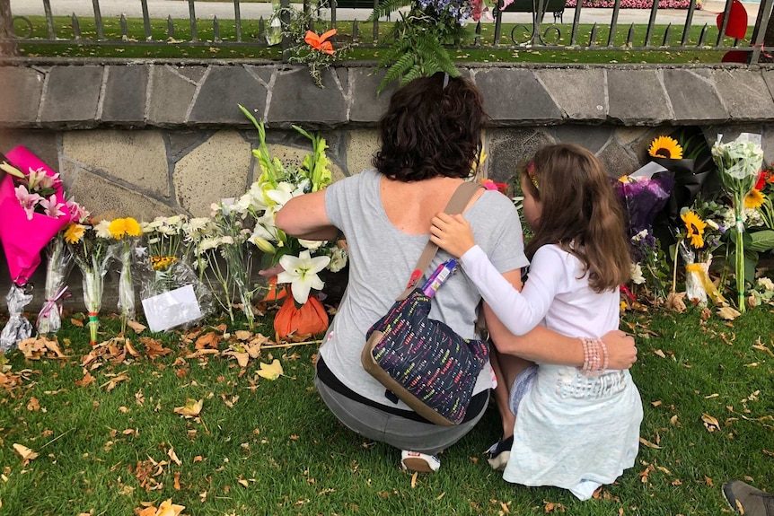 A woman and child kneel in front of a flower covered memorial in the Christchurch Botanic Gardens.