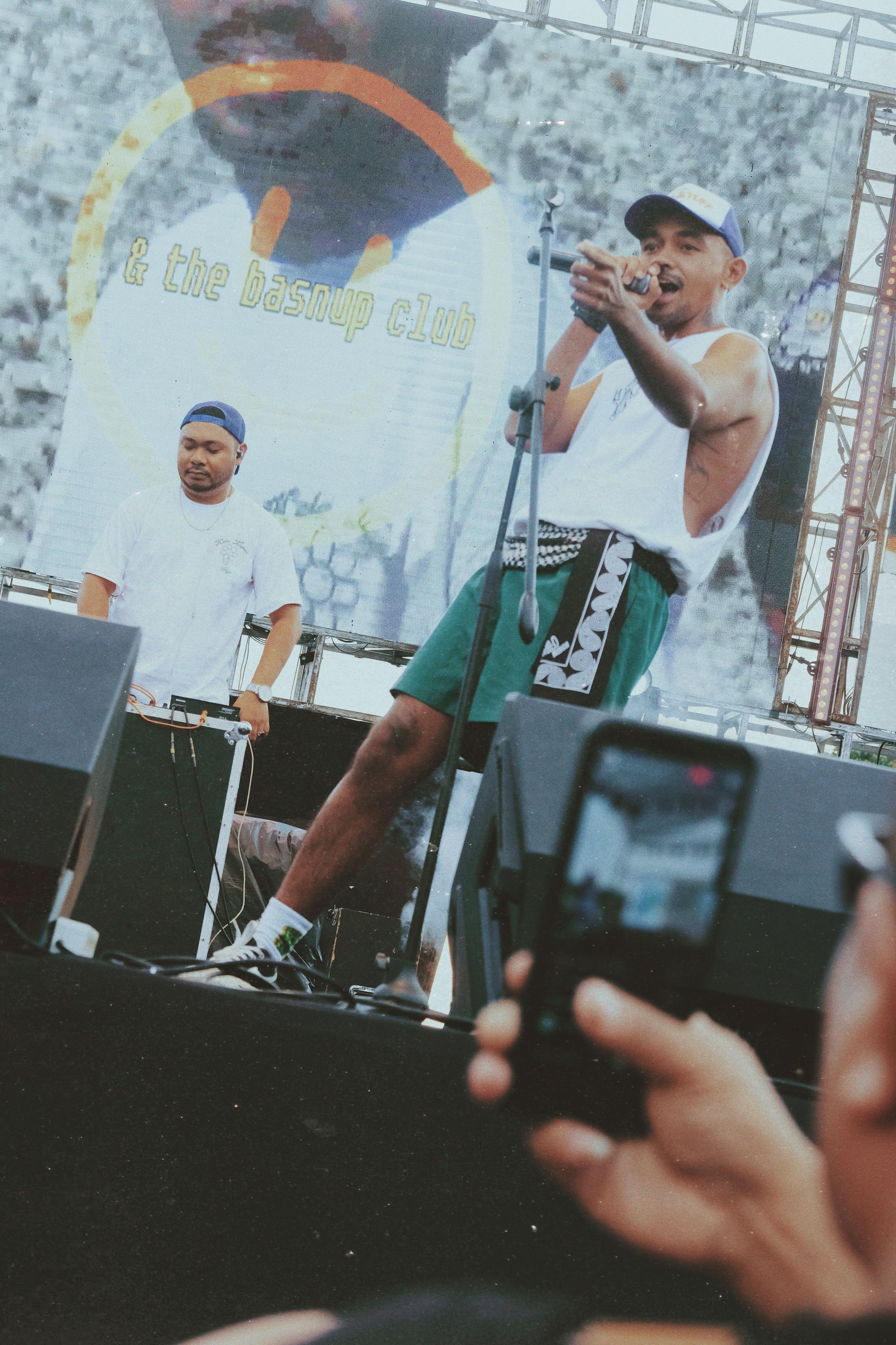 A man in a hat, wearing a white singlet sings on stage, with people taking pictures of him with cellphones in the foreground.