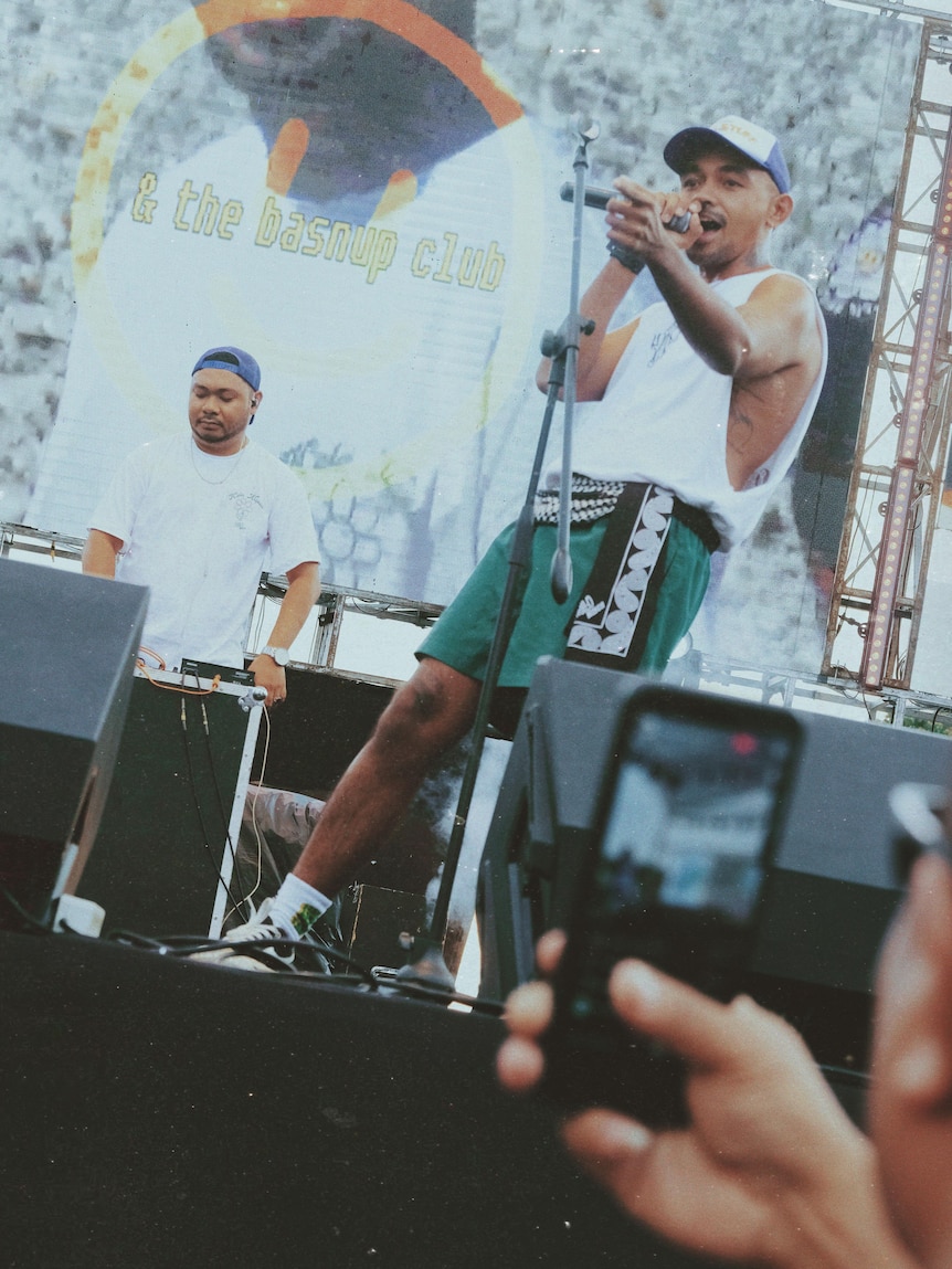 A man in a hat, wearing a white singlet sings on stage, with people taking pictures of him with cellphones in the foreground.