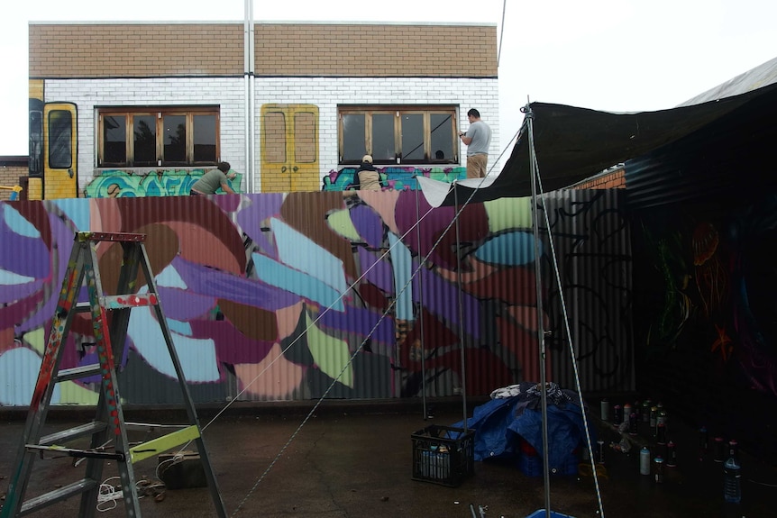 Two men crouch in front a mural on an elevated wall. A ladder and painted wall can be seen in the foreground.