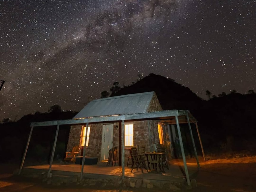 A small stone cottage with light peaking through the windows is beneath a sky full of stars. 