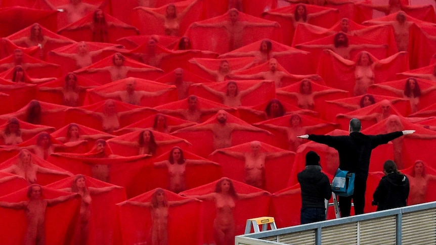 Spencer Tunick directs nude models draped in red fabric to spread their arms out from the shoulders.