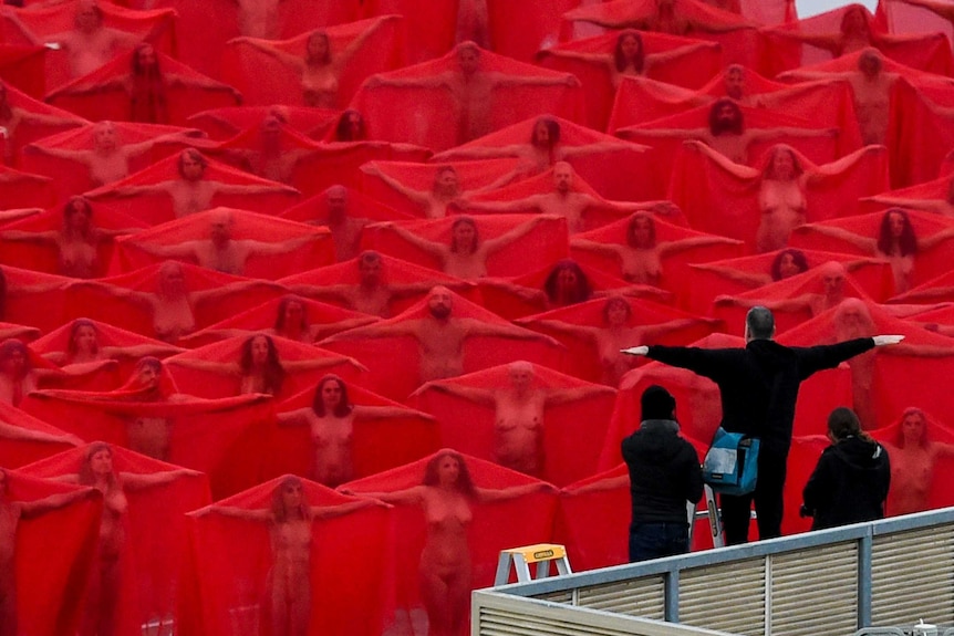 Spencer Tunick directs nude models draped in red fabric to spread their arms out from the shoulders.