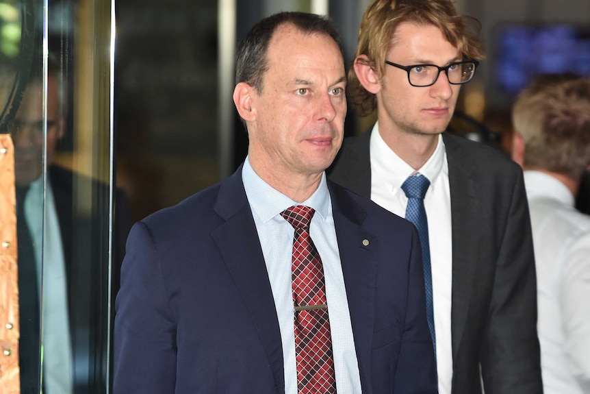 A middle-aged man wearing a blue suit and red checked tie followed by a younger man.