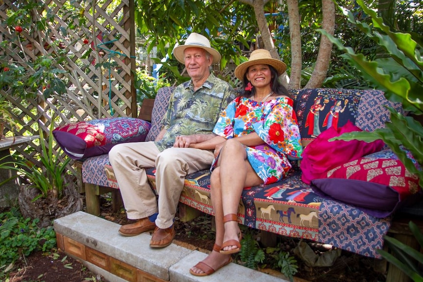 Man and woman sit on brightly coloured couch with green trees and plants around them.