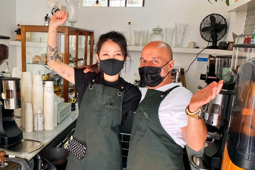 A man and a woman wearing face masks behind a cafe counter raise their arms in celebration.