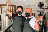 A man and a woman wearing face masks behind a cafe counter raise their arms in celebration.