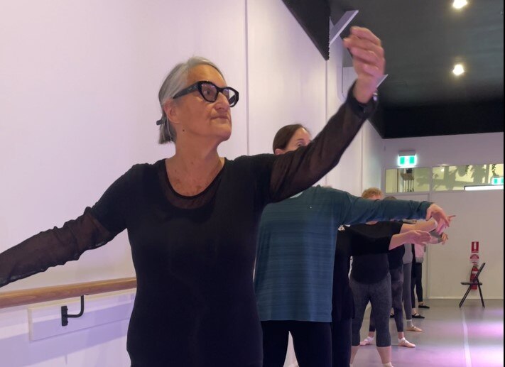 A woman with grey hair pulled back has one hand on the bar and one hand extended in a ballet pose