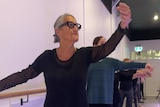 A woman with grey hair pulled back has one hand on the bar and one hand extended in a ballet pose