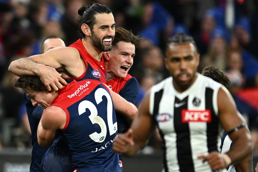 Three AFL players wearing red and blue, celebrate a goal, while an opposition play looks downcast