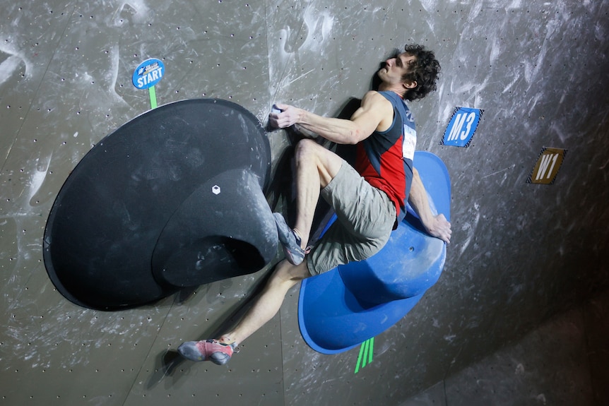 Climber Adam Ondra in an awkward position while bouldering on blue and black holds in a World Cup event.