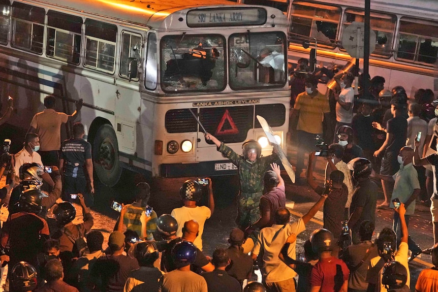 Un policía de Sri Lanka agita un bate en el aire mientras intenta dispersar a los manifestantes frente a un autobús con las ventanas rotas.