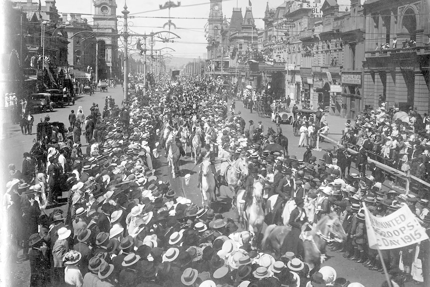 Black and white photo of a large street procession of white horses surrounded by people wearing hats.