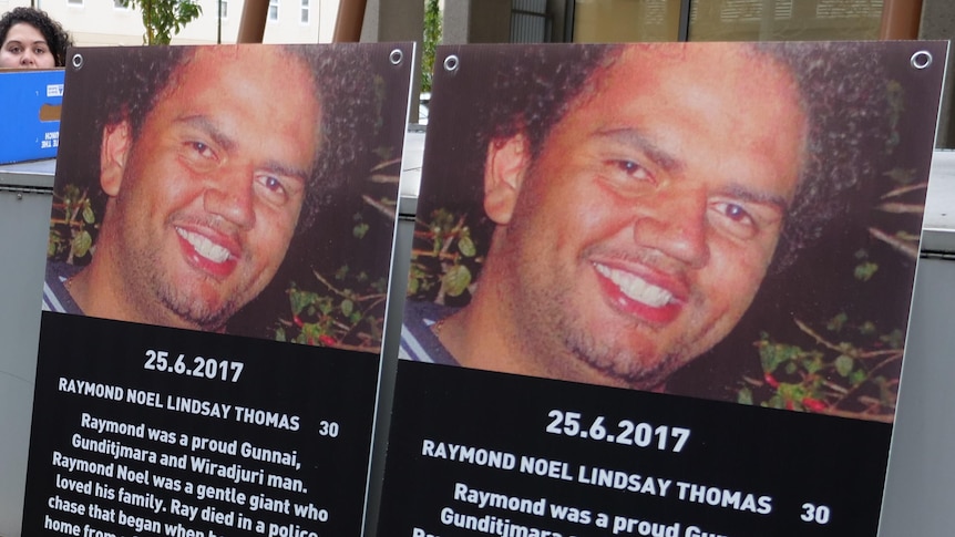 Two posters sit side-by-side on a ledge outside a court showing the face of a man who died and his DOD.  