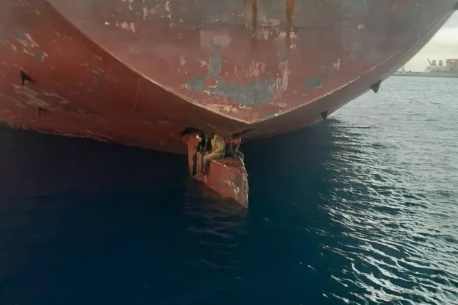 Three men sit on the rudder of a large tanker ship.