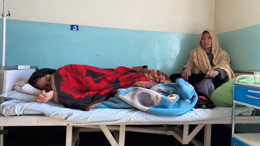 A girl lies in a hospital bed with a red blanket on top of her as a woman watches on.