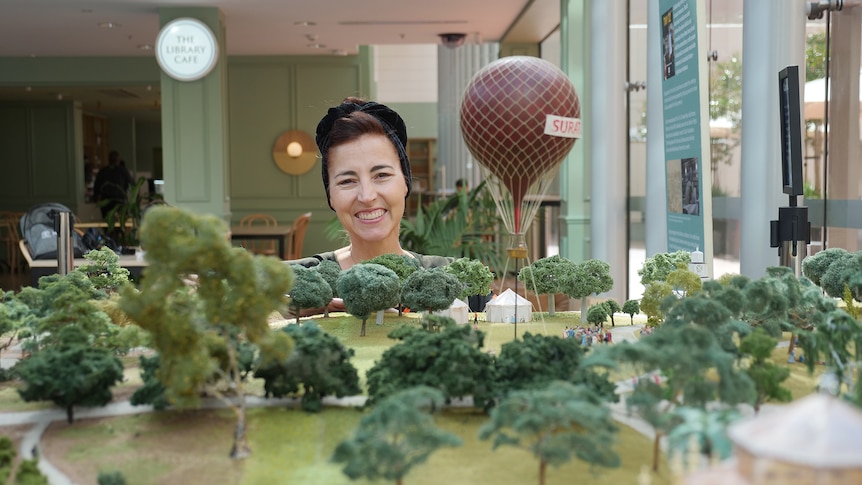 A woman smiles behind a model of a park with trees and a red hot air balloon