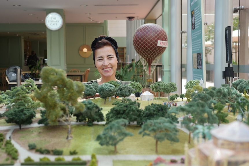 A woman smiles behind a model of a park with trees and a red hot air balloon