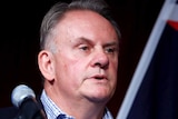 Mark Latham stands behind a microphone. There are two Australian flags behind him.