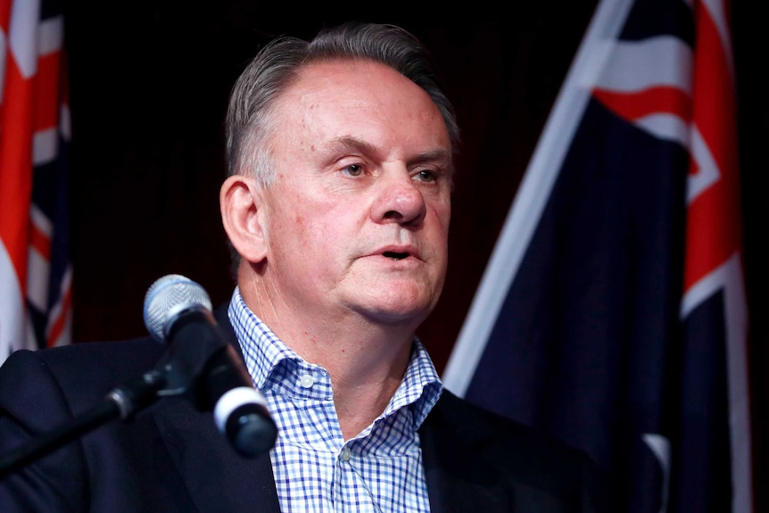 Mark Latham stands behind a microphone. There are two Australian flags behind him.