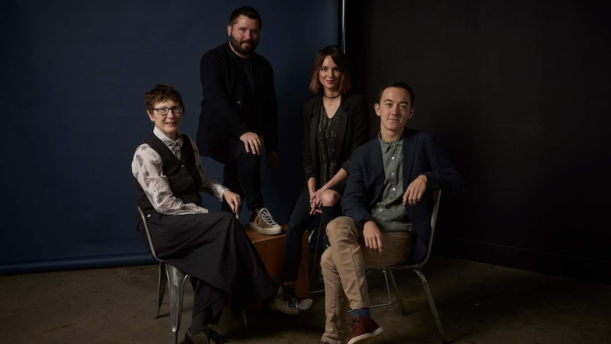 The four theatre makers pose on chairs in front of a moody, navy blue background.