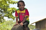 A boy wearing a colourful shirt sitting on a tyre stuck upright out of the ground.