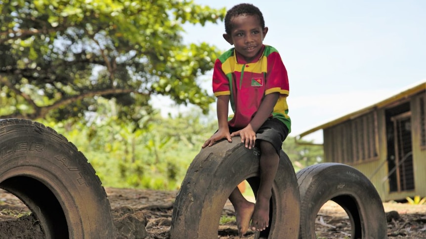 A boy wearing a colourful shirt sitting on a tyre stuck upright out of the ground.
