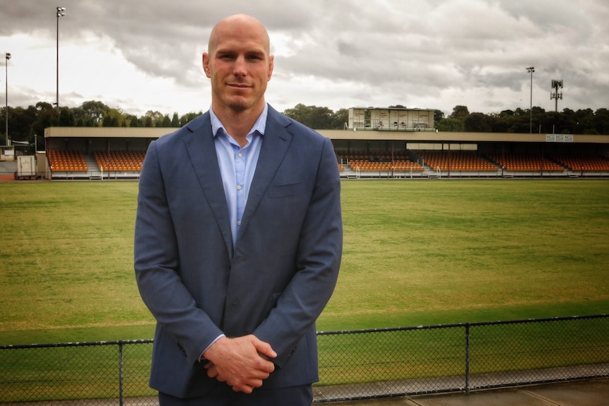 A bald man in a suit stands in front of a sporting field.