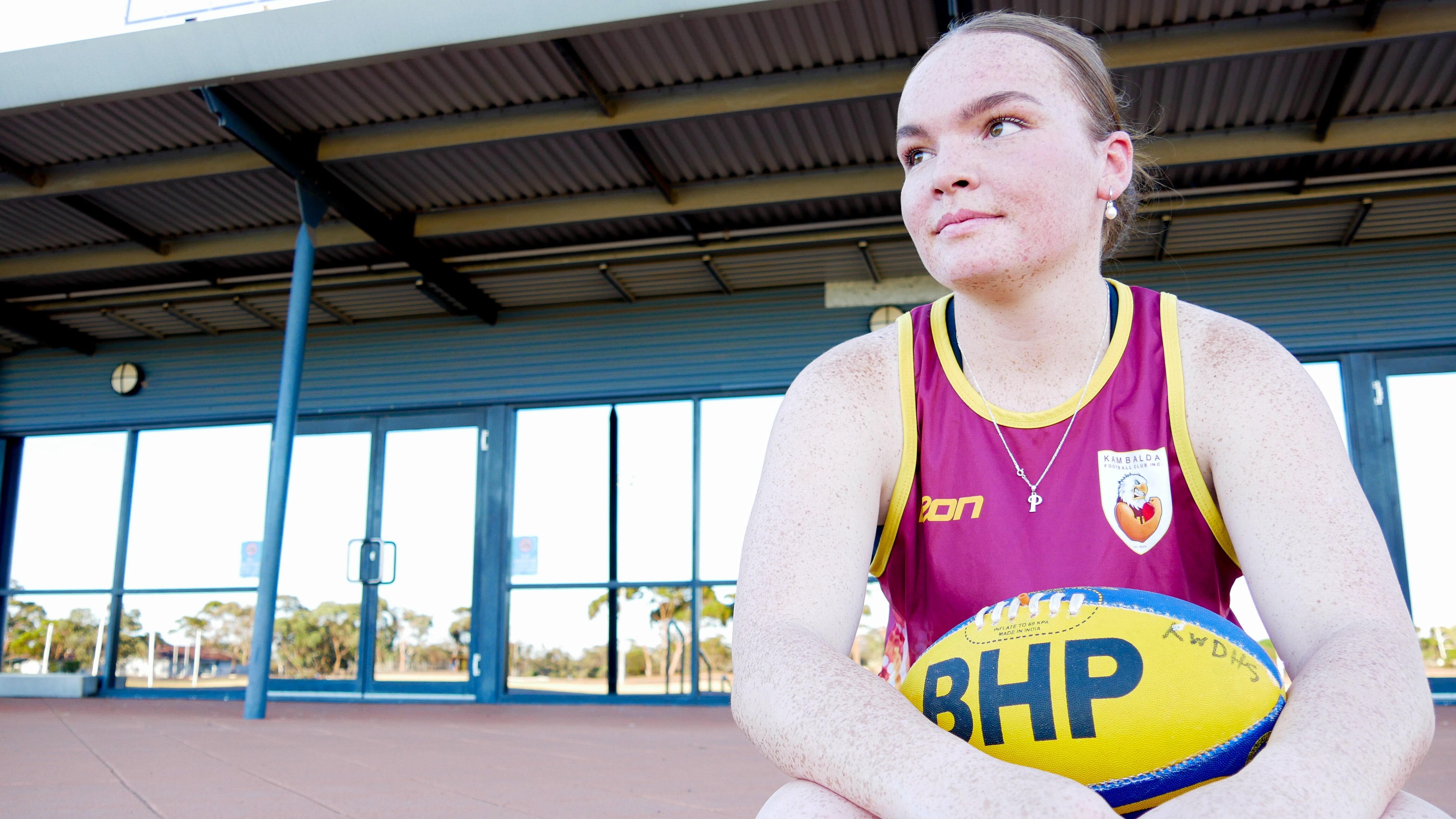 A teenage girl looks off and smiles into the distance holding an Australian rules football, in front of a football club.