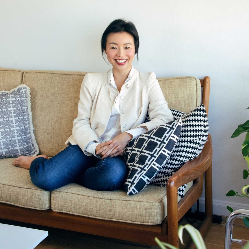 A woman in a white shirt and blazer sits on a couch, she smiles at the camera