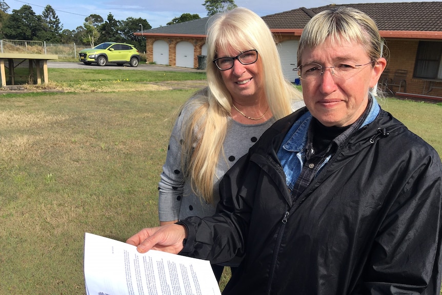 Two blonde, bespectacled women standing outside a brick house.  One holds a document.