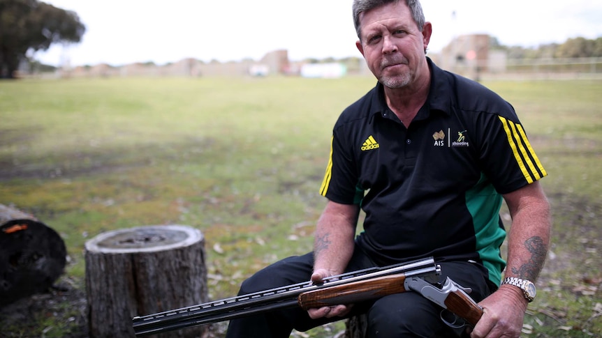 A man in a polo shirt sits on a stump with a cocked gun.